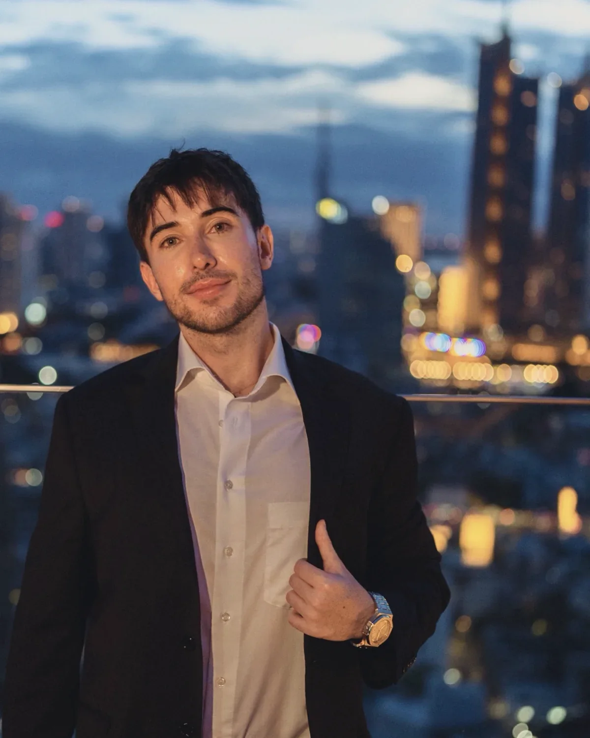Alexander Weiskopf in a dark suit on a rooftop terrace at dusk, the lights of a city behind him